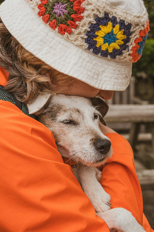 Person in orange jacket hugs a dog wearing a hat.